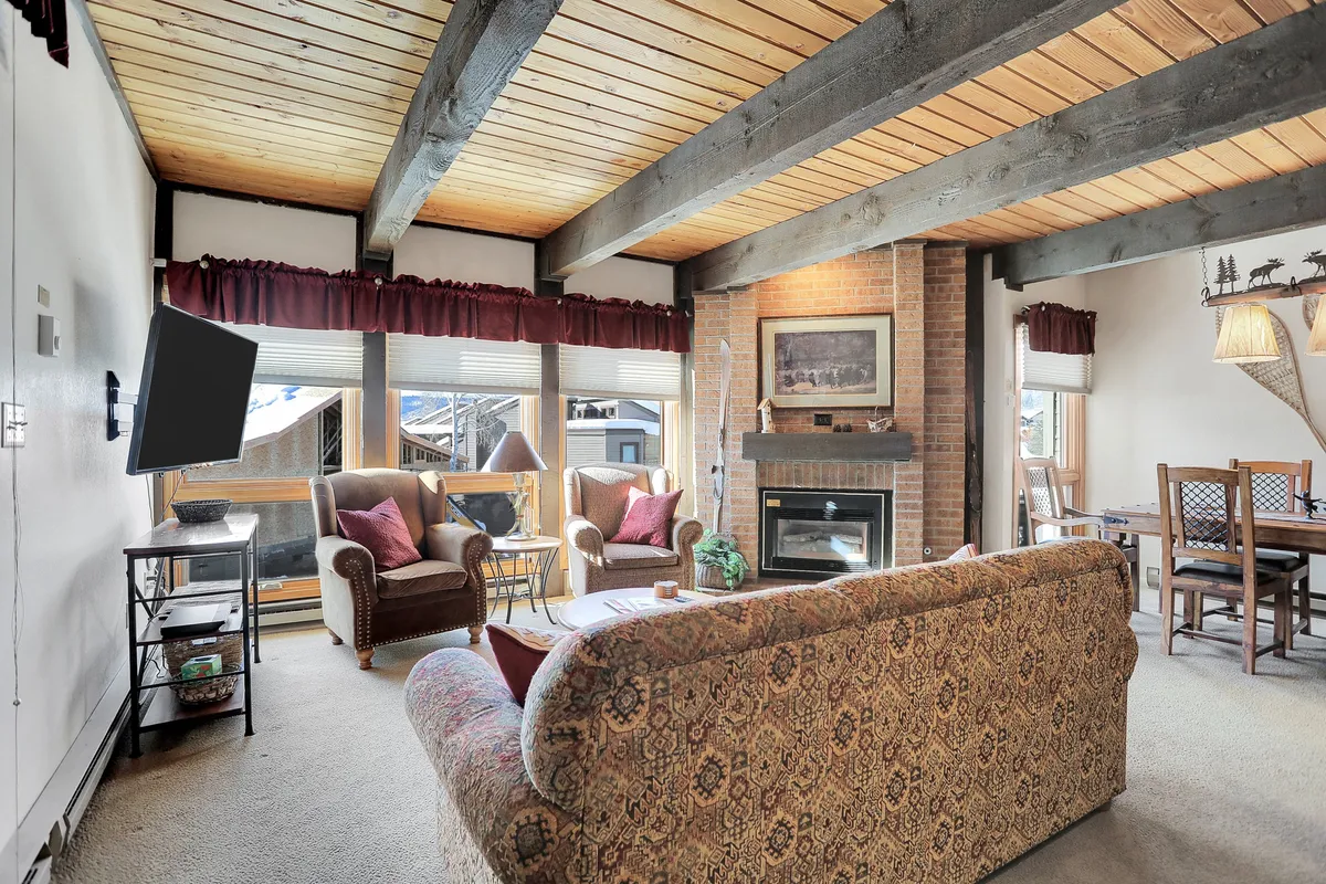 This image features an open living space with a central fireplace, a plush sofa, a dining table, and large windows that let in natural light and offer views of the snowy mountains outside. The scene exudes warmth and comfort, inviting guests to relax, socialize, or enjoy a game night by the fire after a day on the slopes.