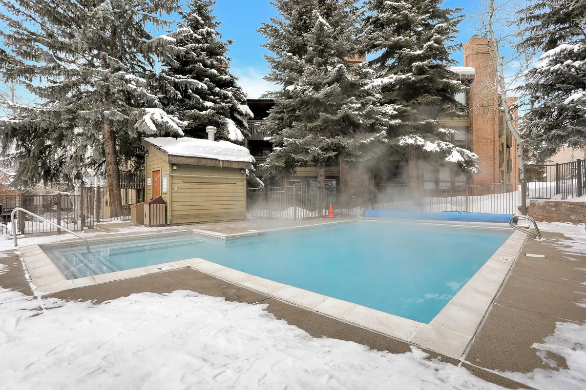 A heated outdoor pool surrounded by snow-covered trees, with steam gently rising from the warm water. The setting is tranquil, with the pool inviting guests to swim or relax even in winter. The crisp blue sky and blanket of snow create a striking resort ambiance.