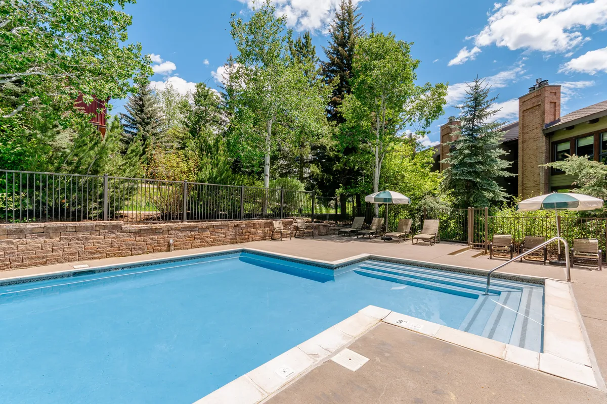 The image showcases the inviting outdoor pool surrounded by lush greenery, comfortable lounge chairs, and umbrellas, creating a relaxing and social atmosphere. The setting highlights spaces ideal for swimming, sunbathing, and gathering with friends or family, all set against the tranquil backdrop of Steamboat Springs.