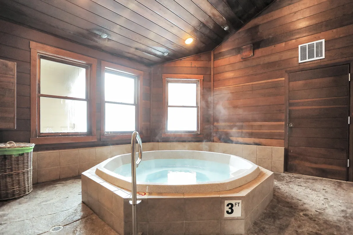 A tranquil indoor setting with a jacuzzi tub beside large windows, wooden walls, and natural light filtering in. The indoor space is serene and private, inviting guests to indulge in soothing soaks regardless of the weather outside.