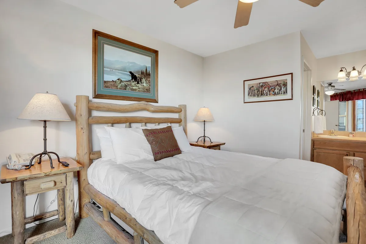 A cozy bedroom with a wooden bed frame dressed in soft linens, complemented by bedside lamps and a decorative painting above the headboard. A vanity area sits nearby, offering a practical yet stylish touch for guests.