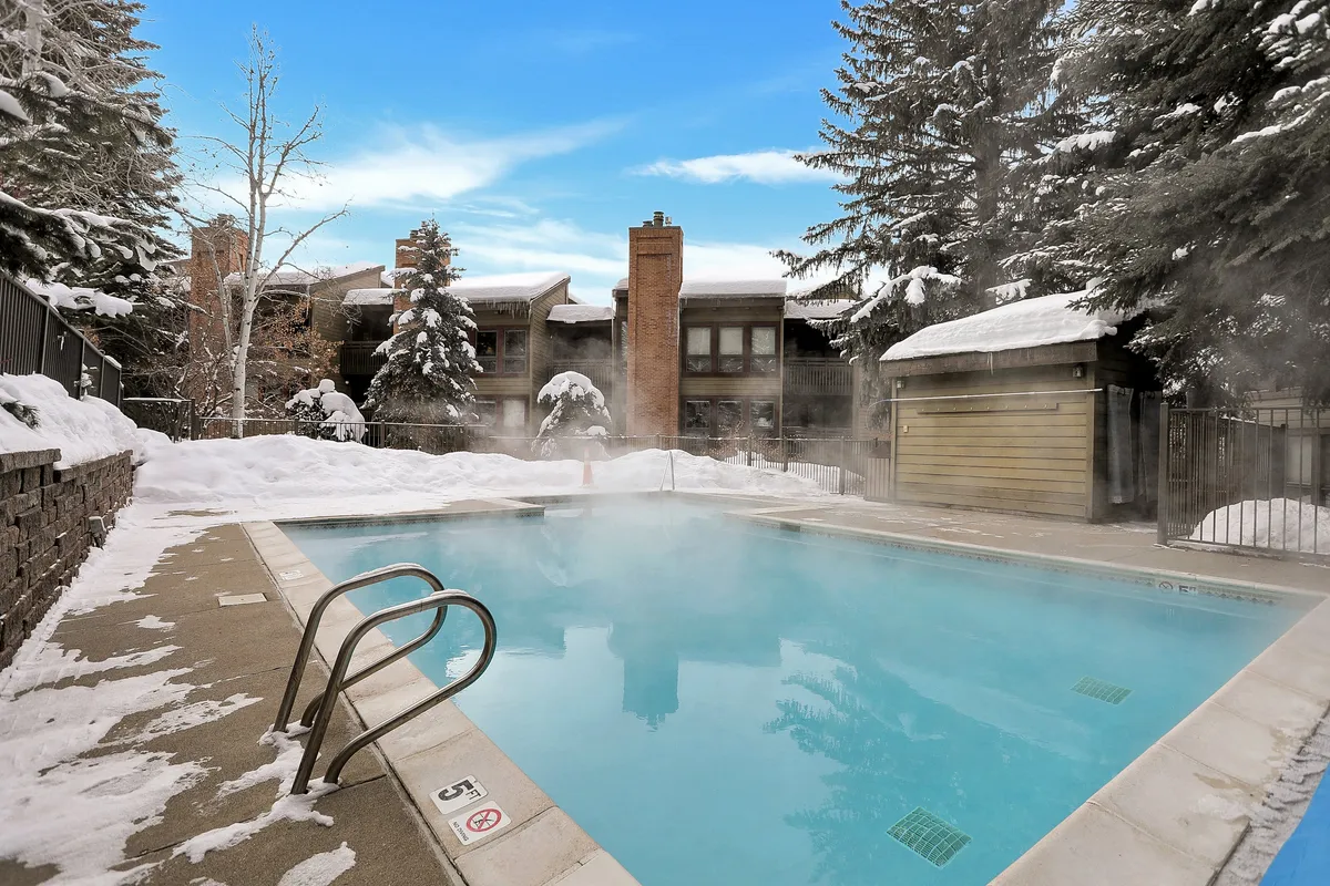 This exterior image displays a heated pool and hot tub set against a snow-covered landscape with tall pine trees and the property in the background. The contrast between the warm water and wintry surroundings evokes a unique, luxurious experience.