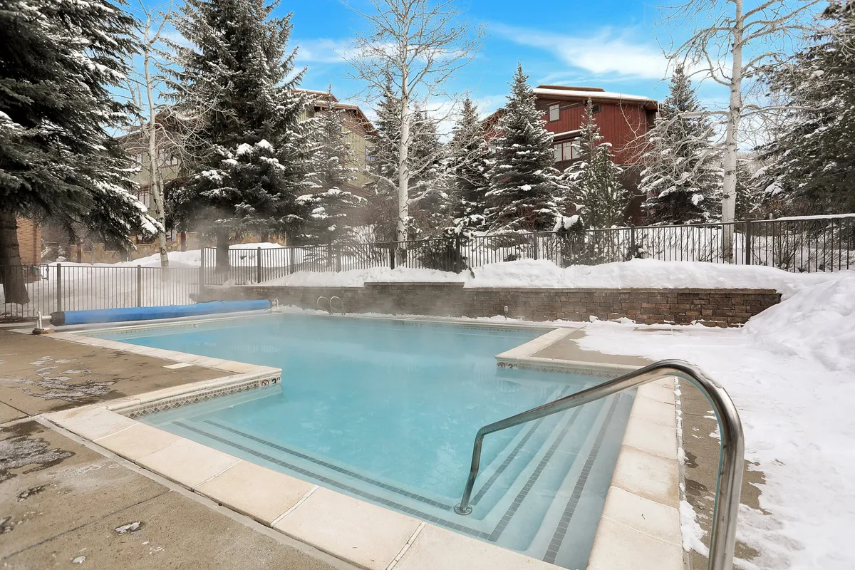 A cozy winter exterior scene featuring a steaming hot tub surrounded by snow and trees, where guests relax and socialize. The setting evokes a quintessential après-ski vibe, perfect for unwinding after dining or brews nearby.