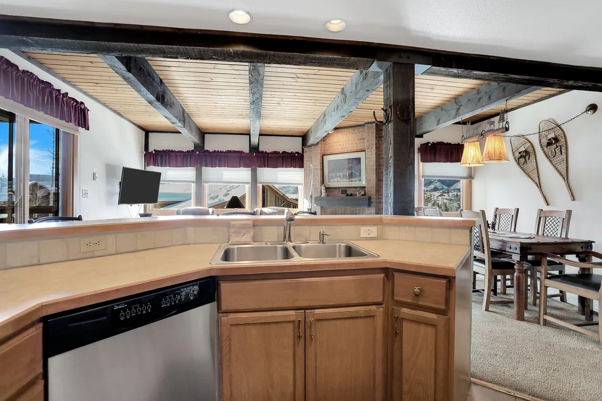 Focusing on the kitchen and adjacent dining area, this image captures the inviting space where cooking, socializing, and sharing meals intersect. Wooden beams add rustic charm, while snowshoes on the wall and a sunlit dining nook suggest a space designed for both culinary creativity and memorable group moments.