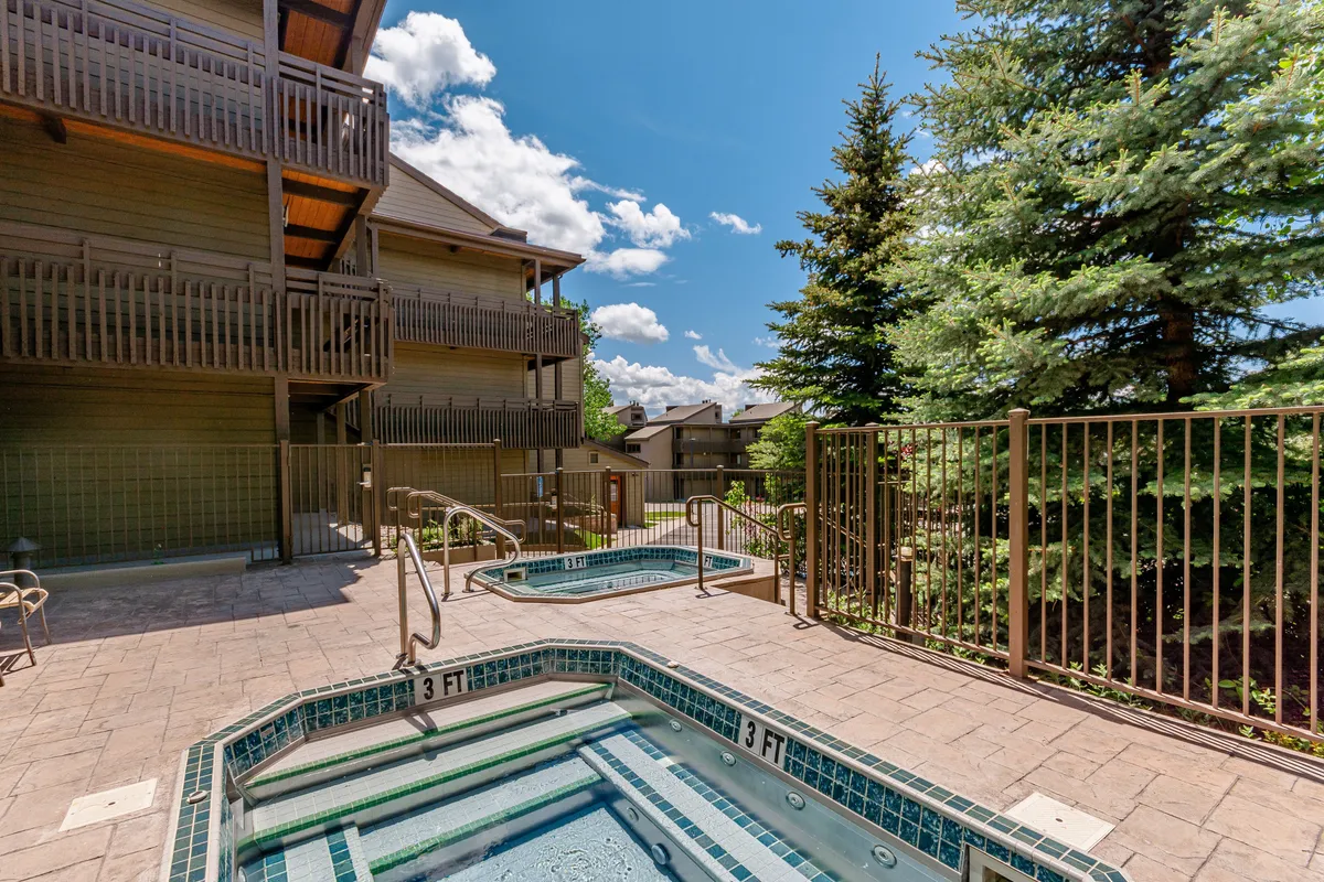 An exterior scene featuring multiple outdoor hot tubs nestled among greenery, with views of the building's architecture in the background. The setting is warm and inviting, ideal for socializing or quiet relaxation at sunset or under the stars.
