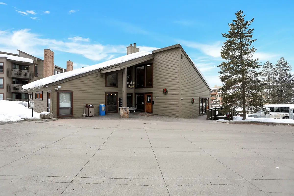 This exterior view captures the building with large windows, a sloped roof, snow-covered ground, and surrounding trees. The entrance area is visible, suggesting a cozy retreat after an active day outdoors.