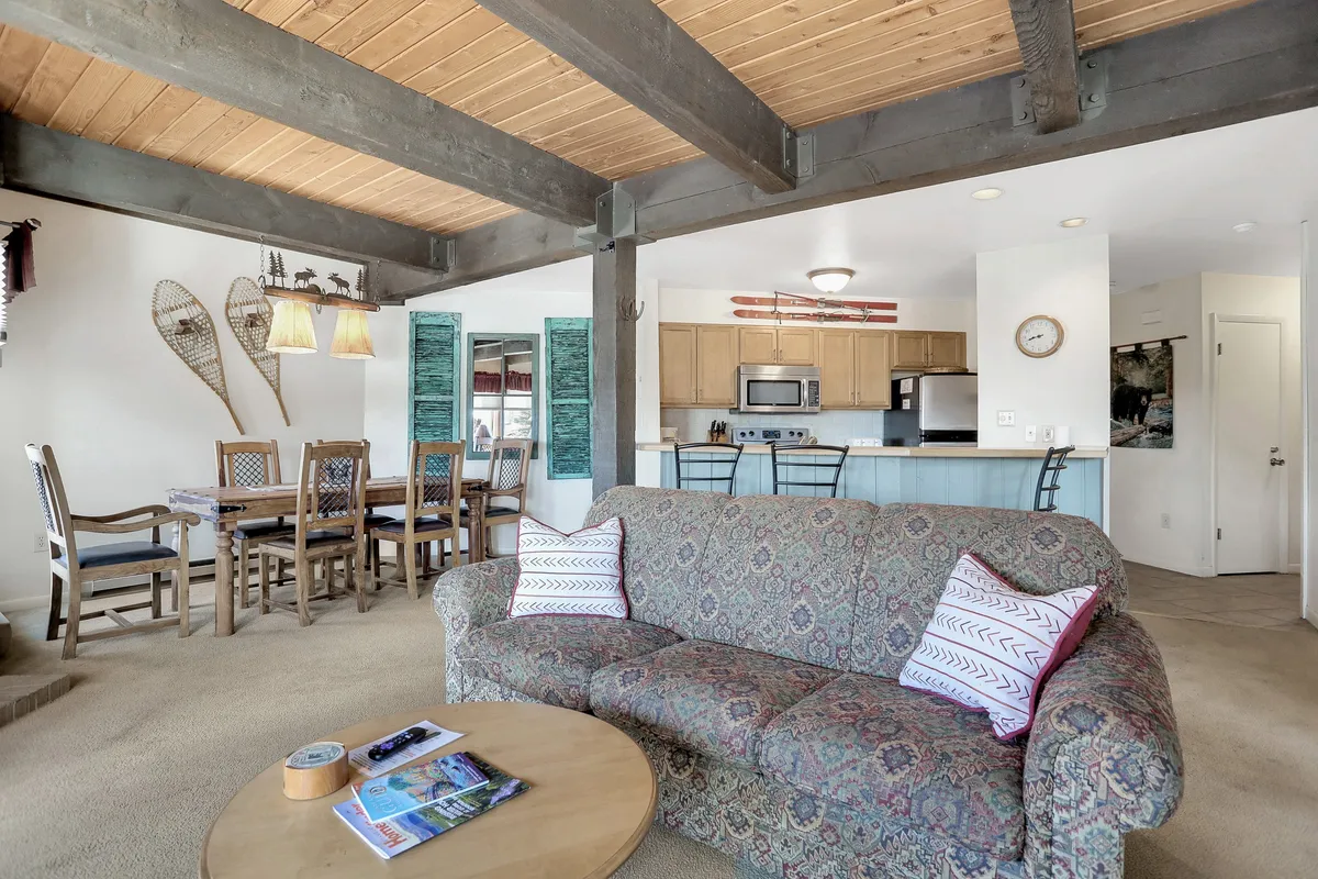 This image showcases a harmonious blend of the living room, dining area, and kitchen. There's a comfortable sofa, a dining table set for a group, and a visible kitchen area with decorative snowshoes and hanging lamps, all under warm, welcoming lighting. The scene suggests togetherness, shared meals, and casual gatherings.