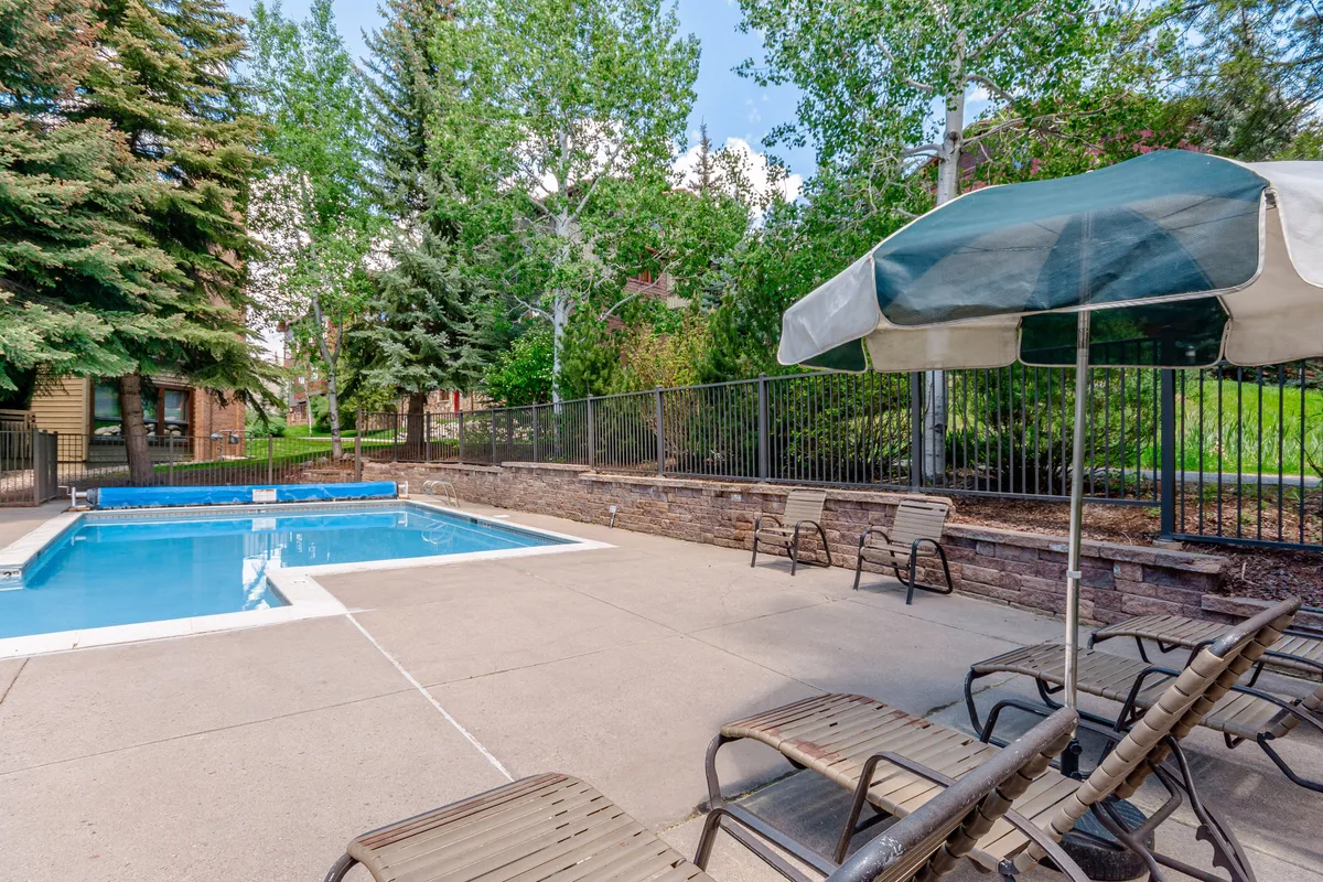 This exterior image showcases a sparkling swimming pool surrounded by lounge chairs, parasols, and lush green trees. The setting is inviting and designed for relaxation and socialization, with ample space for families and groups to unwind by the pool.