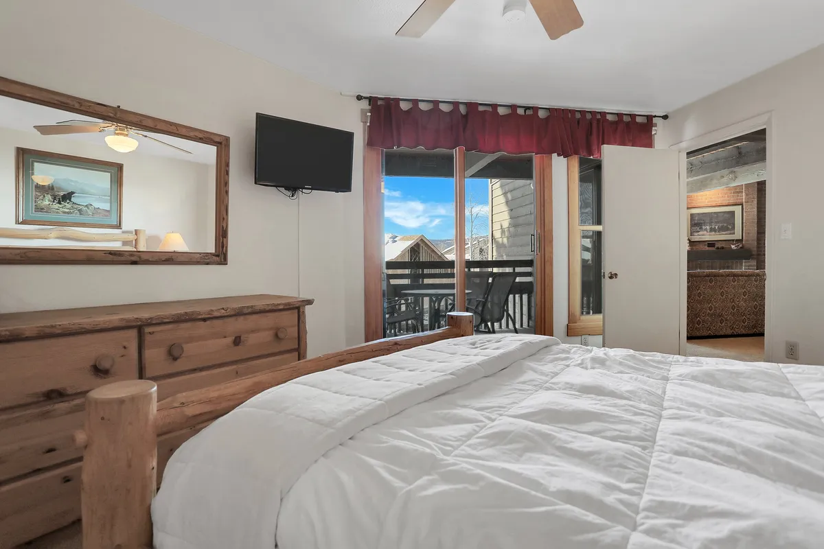 A tranquil bedroom featuring a plush bed with crisp white linens, a wooden dresser, and a TV mounted on the wall. Large glass doors open directly to a balcony, allowing natural light to fill the room and providing an inviting spot for morning coffee or evening relaxation.